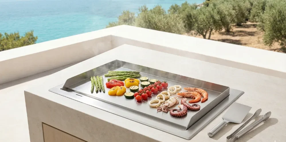 Overhead view of a stainless steel Teppanyaki griddle integrated into an outdoor kitchen, steaming shrimp, octopus, and vegetables with views of the Mediterranean sea.