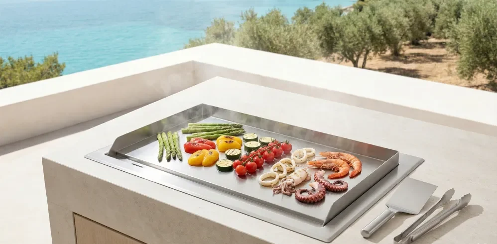 Vista superior de una plancha Teppanyaki de acero inoxidable integrada en una cocina exterior, cocinando gambas, pulpo y verduras al vapor con vistas al mar Mediterráneo.